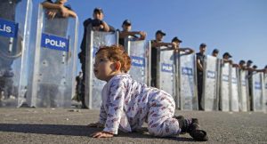epa04937946 A Syrian refugee baby plays in front of riot police at the Istanbul-Edirne highway as they wait for permission to pass Turkish Greek border to reach Germany in Edirne, Turkey 19 September 2015. Turkey has spent 7.6 billion US dollar on caring for 2.2 million Syrian refugees since the civil war began in 2011, Deputy Prime Minister Numan Kurtulmus said on 18 September. Turkey hosts more Syrians who fled their homeland than any other country. Most Syrians, however, have no legal right to work and rights groups report housing remains a huge issue for many. EPA/TOLGA BOZOGLU EPA/TOLGA BOZOGLU